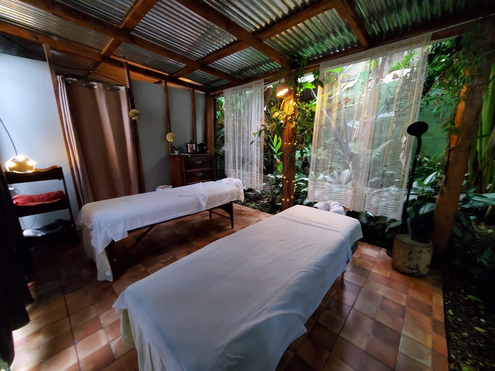 Sala de masajes para parejas al aire libre, con techo de zinc, vigas de madera y cortinas
